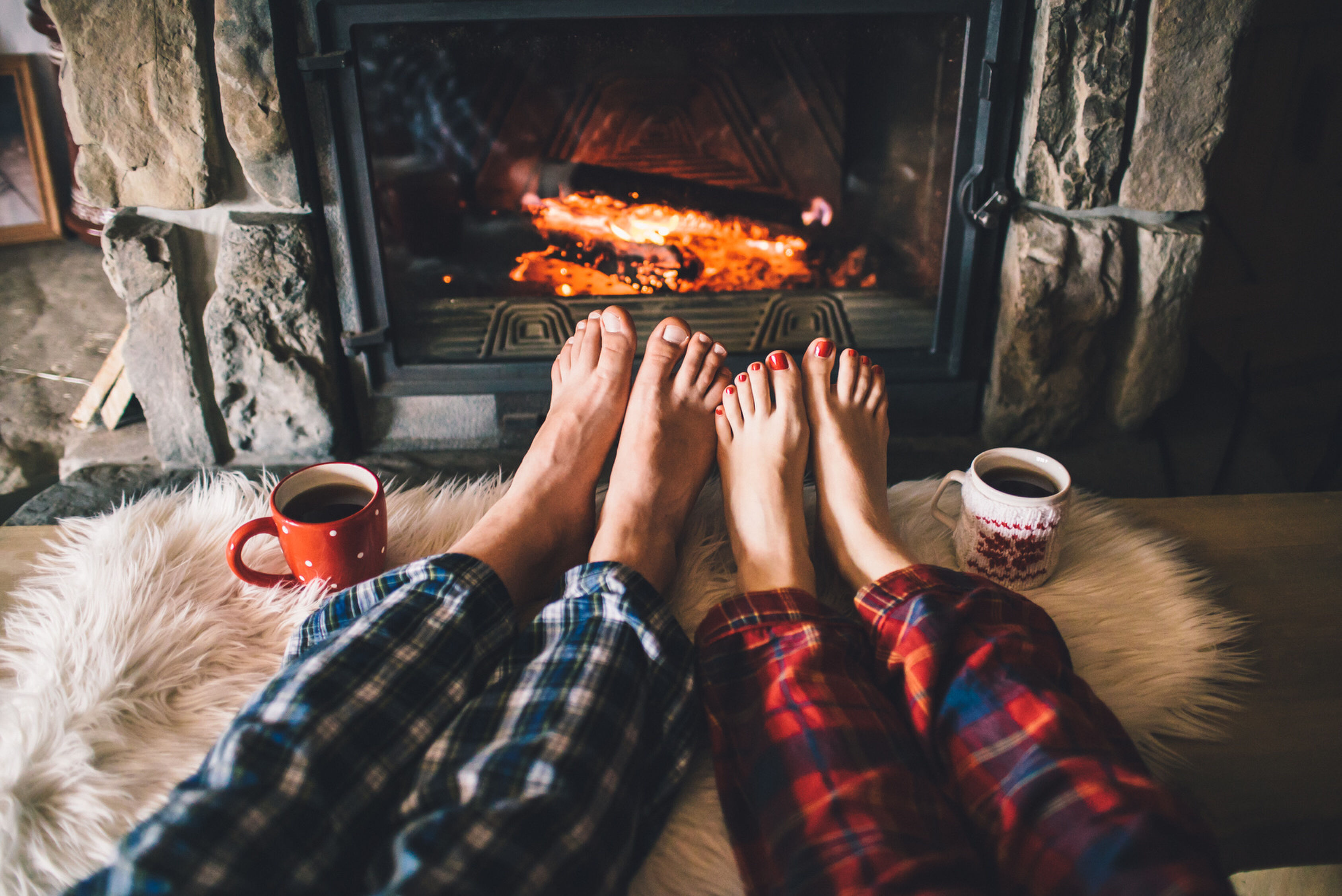 Bare couple feet by the cozy fireplace. Man and Woman relaxes by warm fire with a cup of hot drink and warming up her feet. Close up on feet. Winter and Christmas holidays concept.