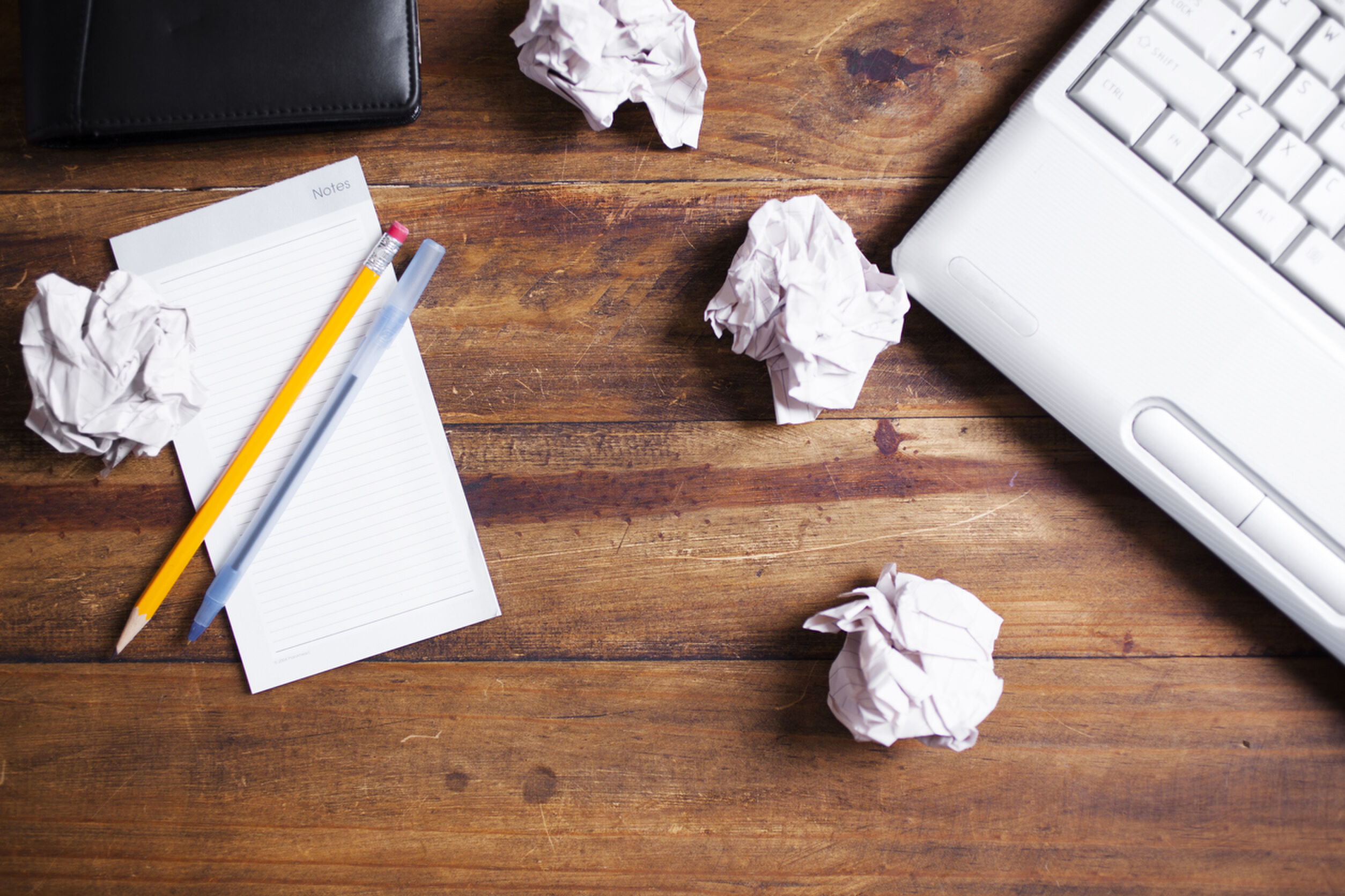 Laptop computer, crumpled paper balls, notepad, organizer all lie on an office desk. Wooden background. No people.