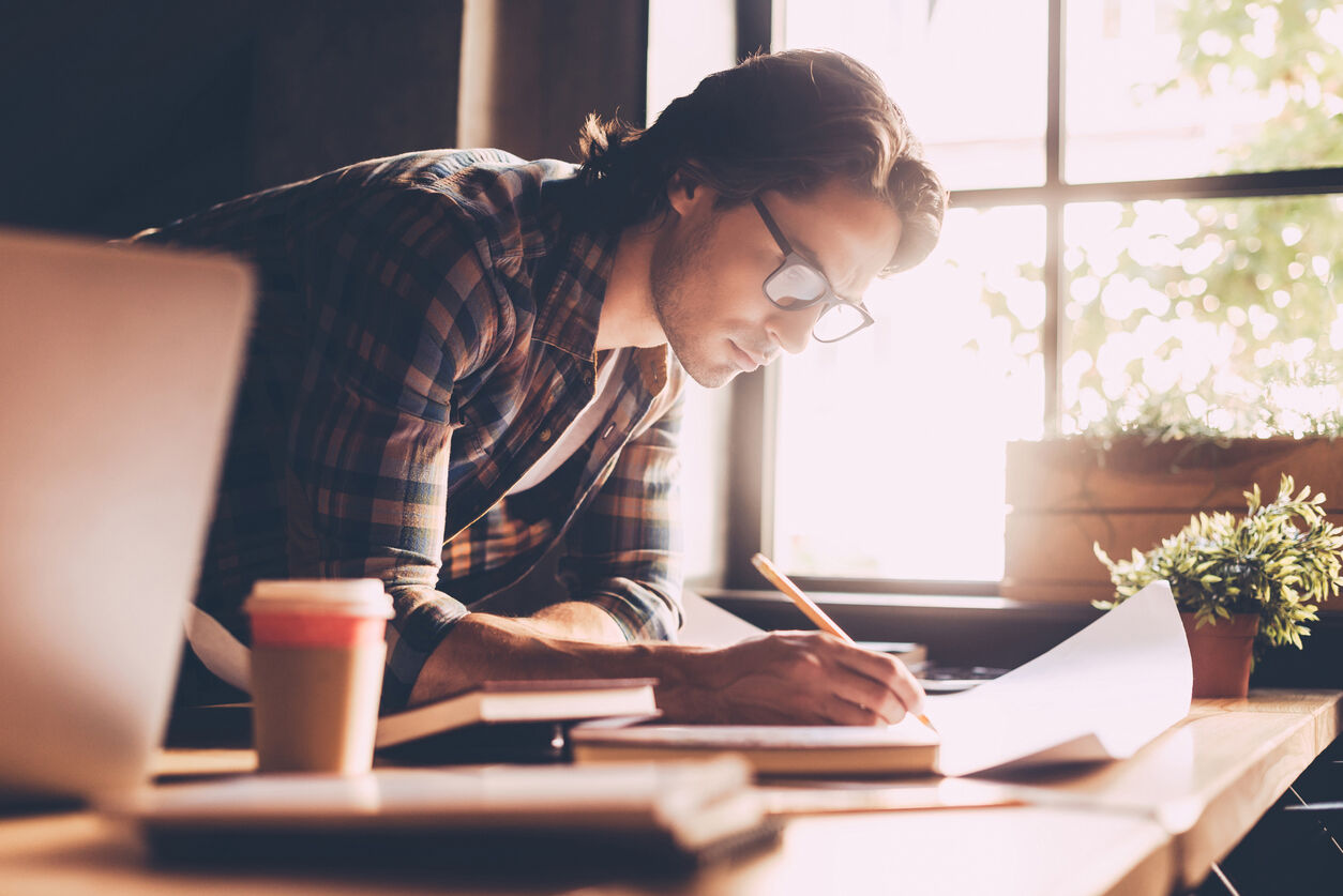 Confident young man in casual wear sketching on blueprint while standing near wooden desk in creative office