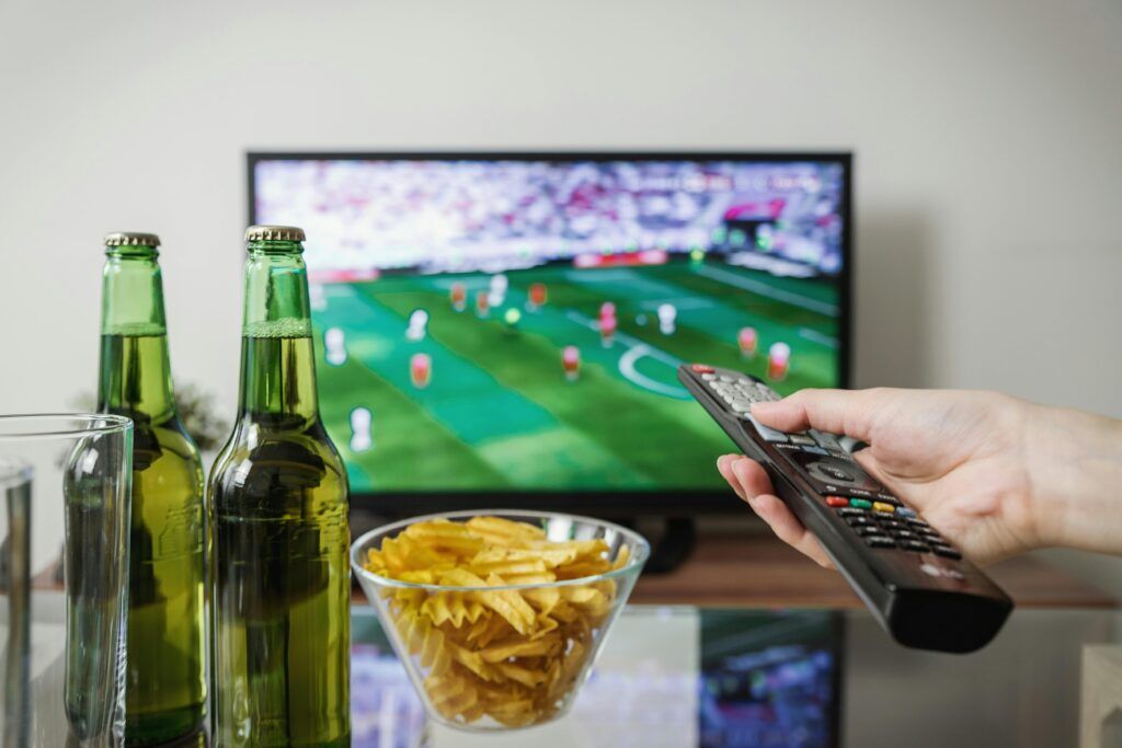 een man is lekker op z'n gemakje tv aan het kijken onder het genot van een chipje en een tweetal biertjes