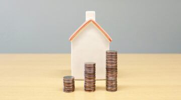 Een huisje met stapeltjes geld ervoor.