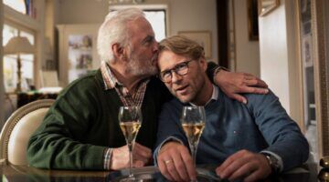 Twee mannen met een glas champagne en de ene man geeft de ander een zoen op zijn hoofd.